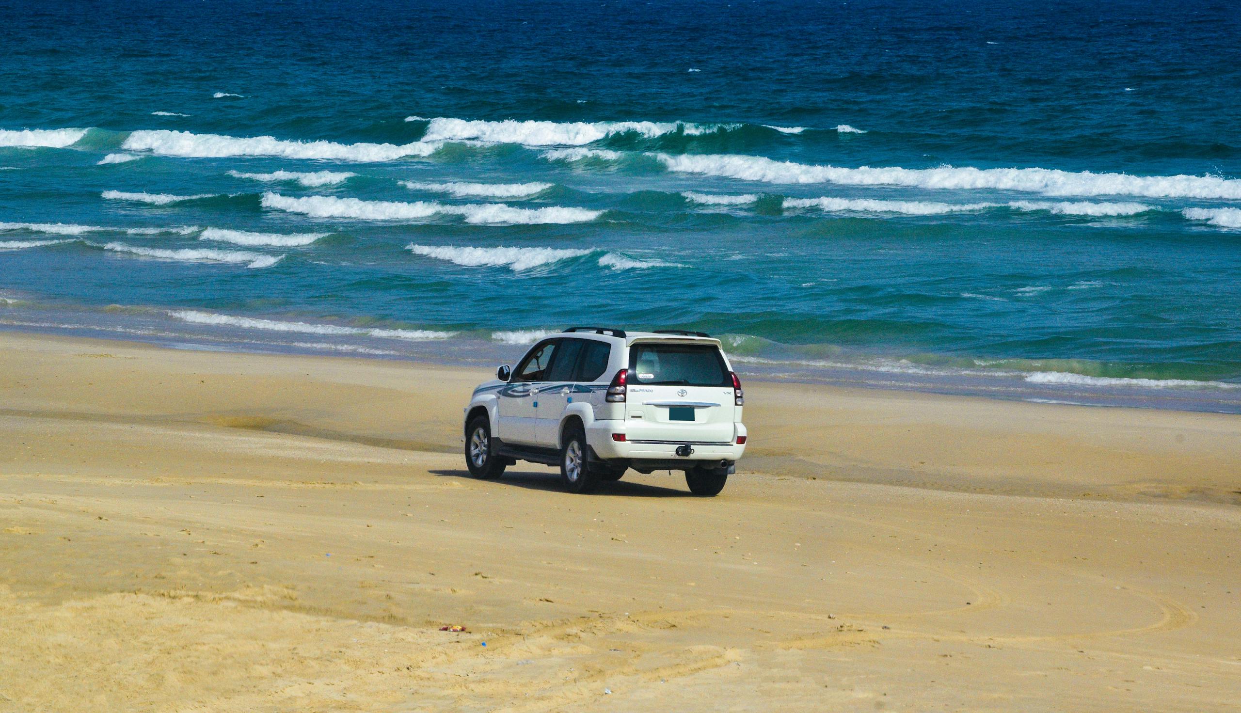 A white SUV traverses a picturesque sandy beach by the ocean under clear skies.