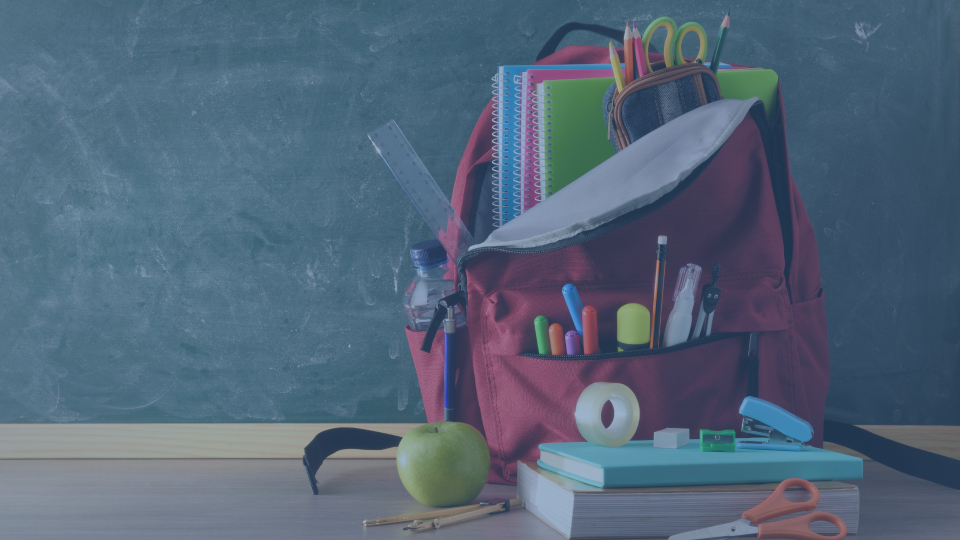 Back-to-school supplies including a red backpack, notebooks, pencils, scissors, and stationery on a desk against a classroom chalkboard.